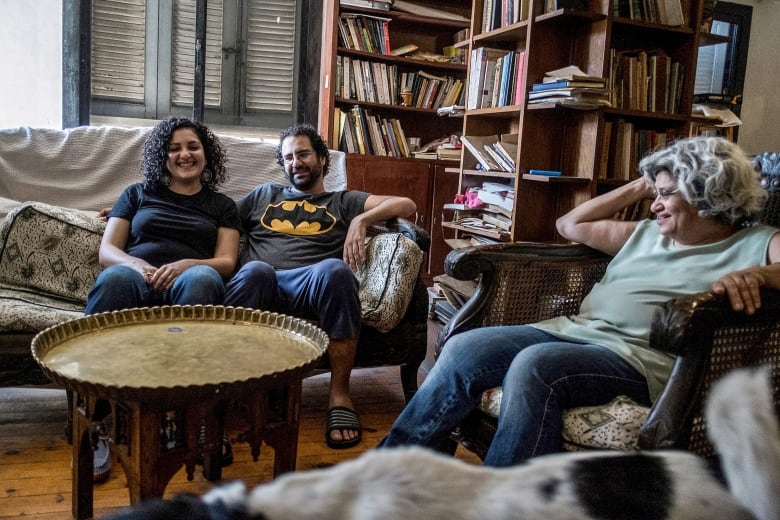 A man and a woman sit side-by-side on a couch smiling. A smiling older woman sits in a chair next to them. They are in a living room filled with bookshelves. 