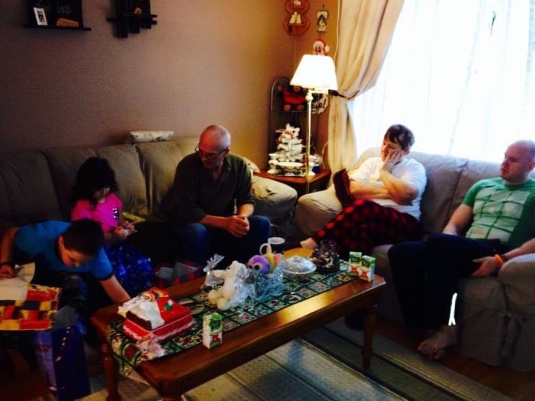 Five people sit on sofas around a coffee table. On the left, two children are unwrapping gifts.