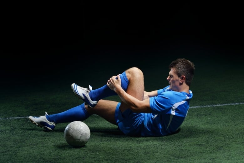 A man in a soccer uniform lies on the grass clutching his knee and scrunching his face in pain.