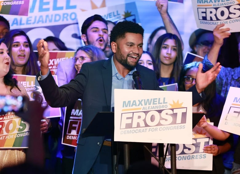 Dark haired individual wearing a blue shirt and jacket, smiling, arm raised, with supporters behind him. 