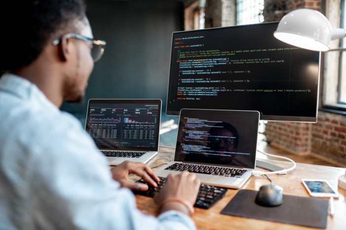 Young African male programmer writing program code sitting at the workplace with three monitors in the office