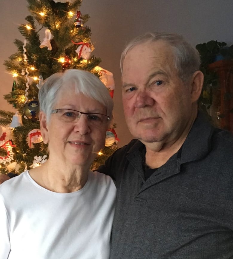 An older couple are smiling into the camera. They're standing in front of a Christmas tree.