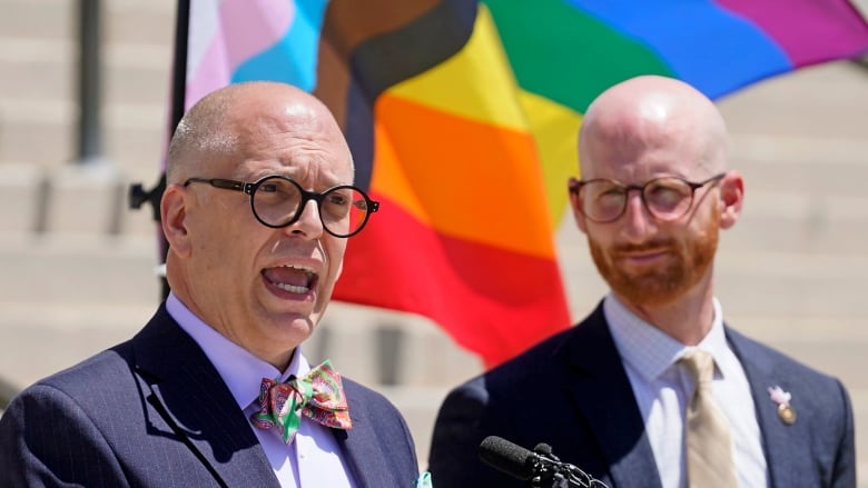 Two nearly bald individuals, both wearing glasses, speak in front of the rainbow flag.