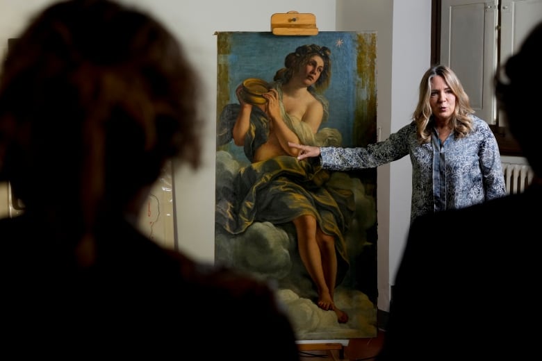 A woman with long blond hair speaks as she points at a large painting of a naked woman holding a pot, her nudity strategically covered by a long, flowing piece of fabric. In the foreground of the photograph are silhouettes of people crowded around watching the woman speak. 