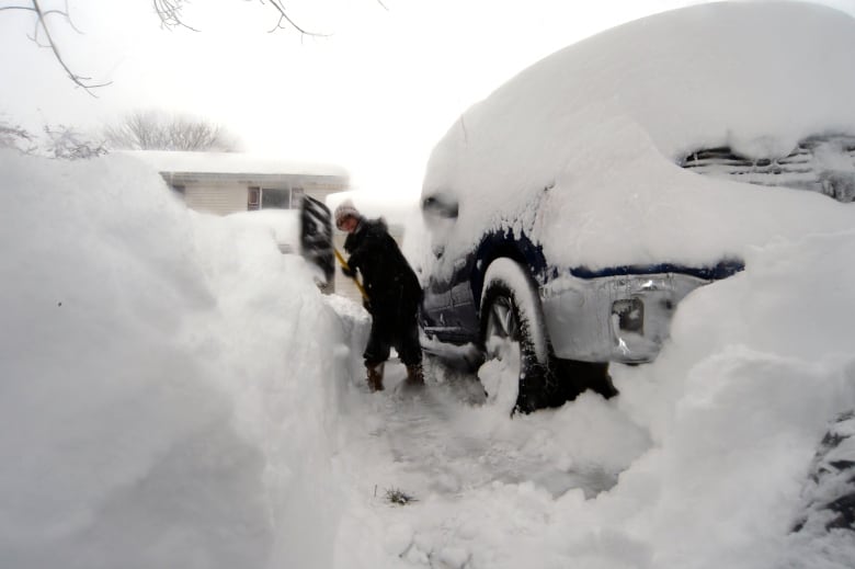 A woman shovels a path alongside a vehicle through deep snow.