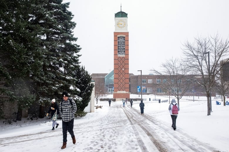 People walk across a snowy college campus. There is a large clock tower in the background. 