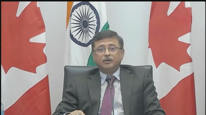 A Zoom screenshot of an Indian man in a suit framed by an Indian and two Canadian flags.