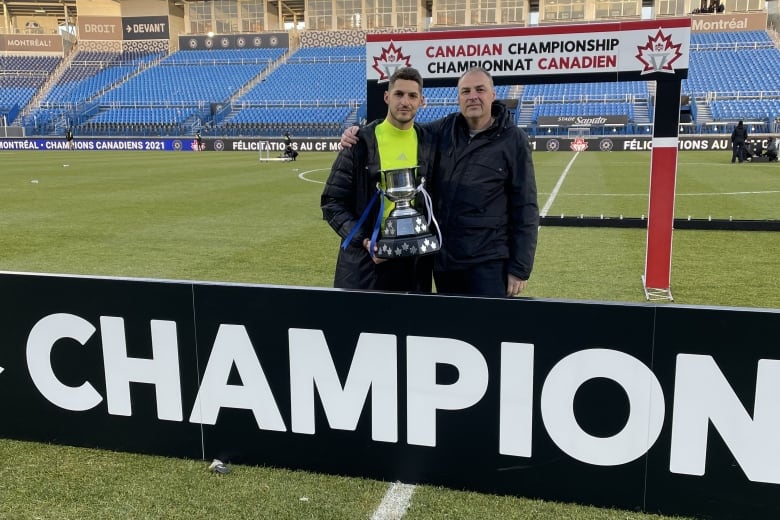 Two men hold a trophy on a soccer field. 