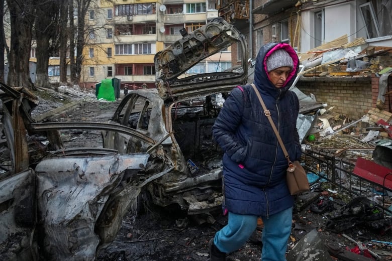 A person bundled in a winter coat and hood walks by a bombed out car and home.