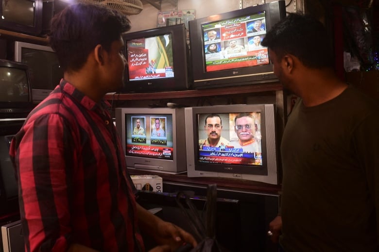 Two men stand watching several televisions showing Pakistani news broadcasts.