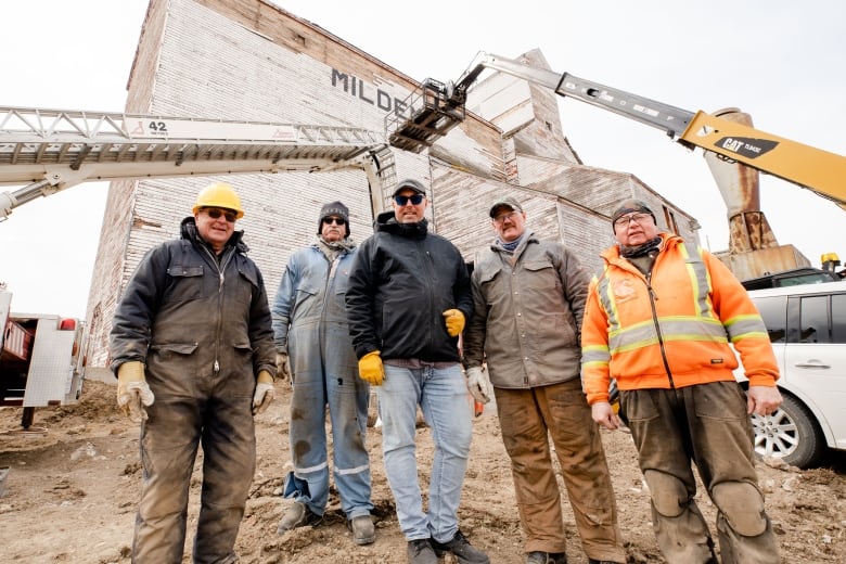 Five men in work clothing stand in front of a wooden grain elevator and ladder trucks.