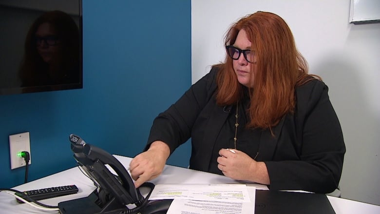 A white woman with straight auburn hair wearing glasses and a black blazer--Go Public reporter Carolyn Dunn--  sits at a white desk pressing the key pad of a desk phone in front of her.