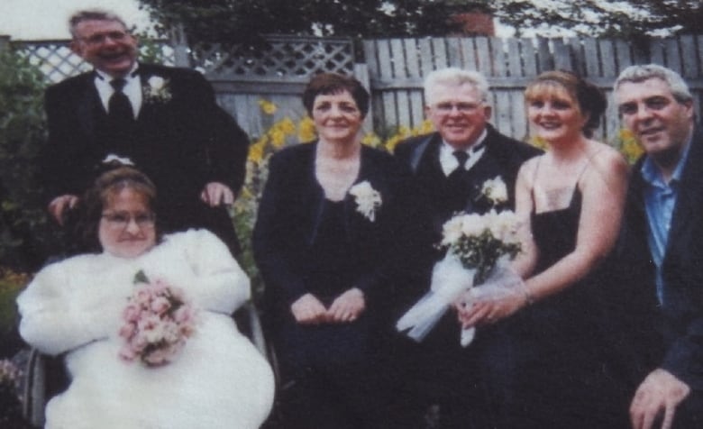 A photo of six people, all facing the camera and smiling. On the far left is a woman in a wheelchair, wearing a wedding dress and holding a bouquet. A man wearing suit and tie stands behind her. Next to them on the right, four people are sitting next to each other, all wearing formal attire.