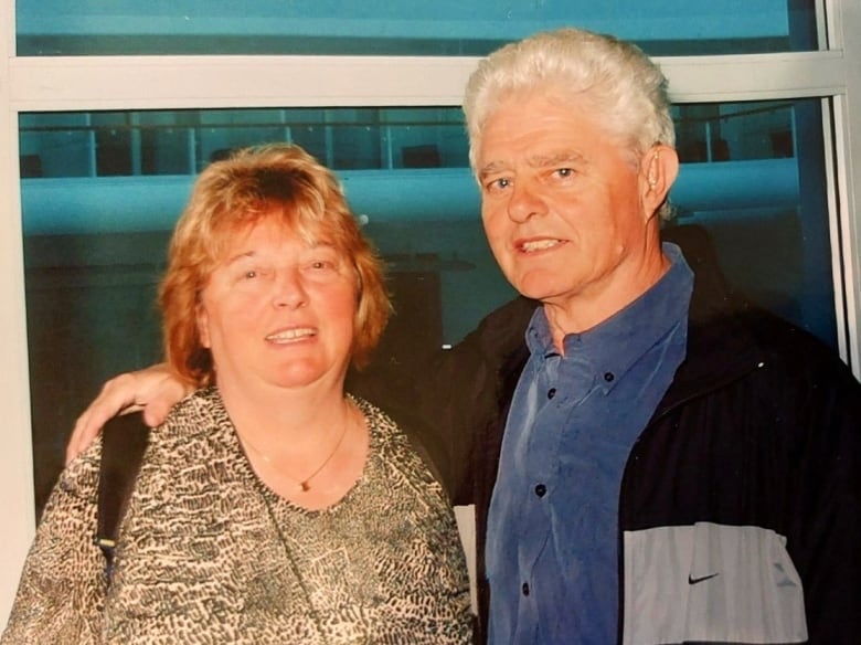 A middle-aged woman, left, and man, right, smile into the camera. He has his right arm around her shoulders.