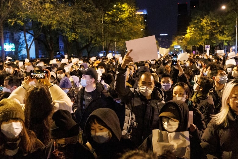 Hundreds of people gathered together outside at night. Some hold up blank white sheets of paper.