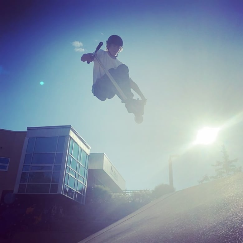 A teenage boy his seen doing a trick on his scooter at a skatepark while the sun is shining.