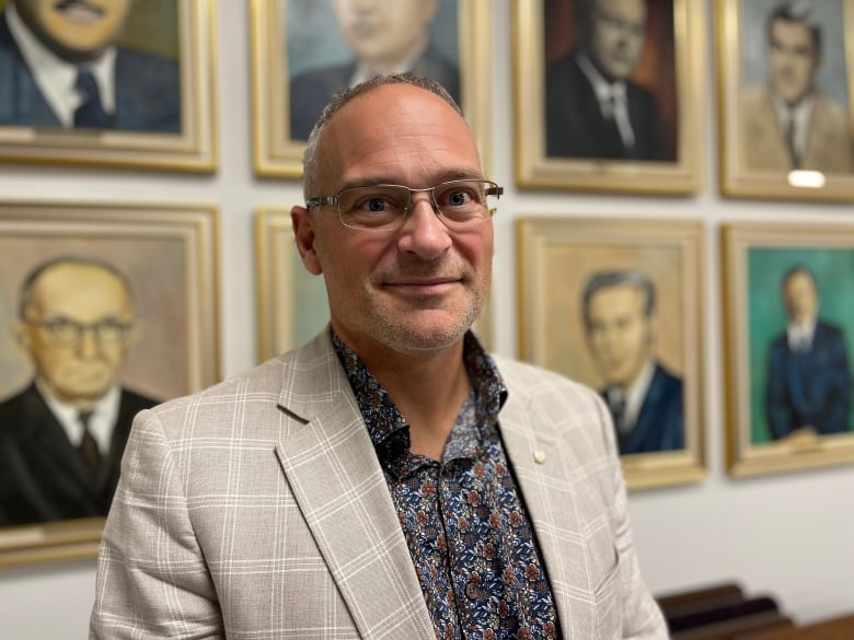 A man with glasses in a pinstripe suit and floral shirt looks straight at the camera. Portraits of former mayors of Amos, Que. are visible behind him.