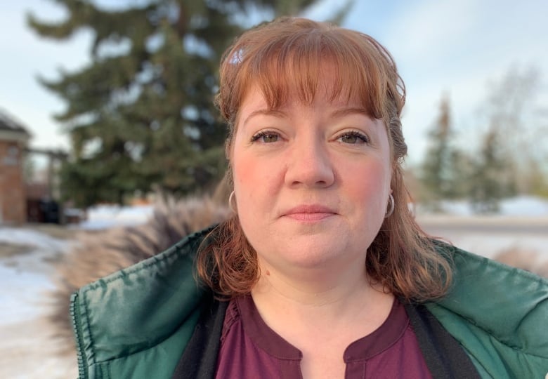 A woman in a green parka stands outside with snow banks and evergreen trees seen behind her.  