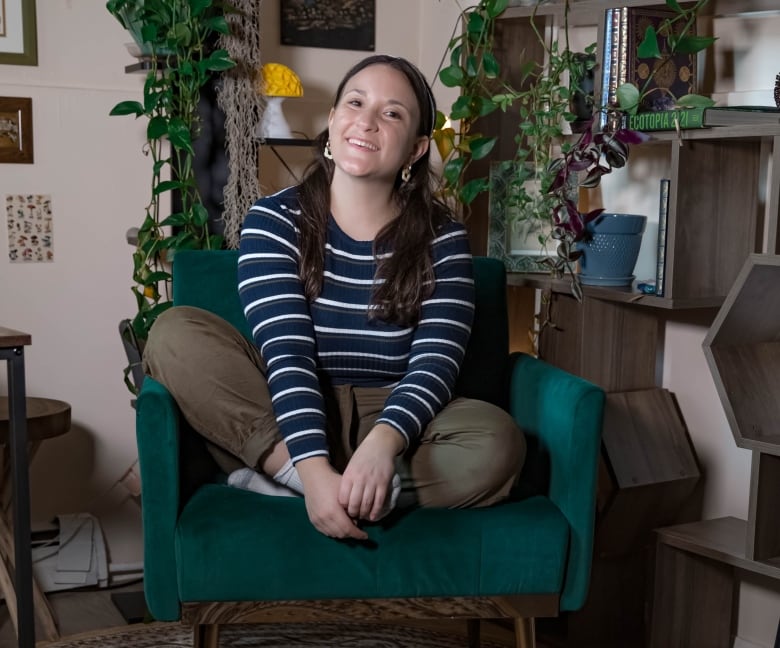 A smiling woman sits crossed-legged in an upholstered teal chair, with a collection of hanging houseplants displayed on a geometrically shaped bookcase behind her.