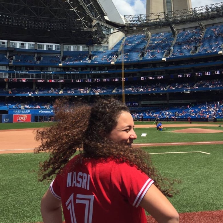 A woman wearing a jersey with the name “Nasri” smiles as she stands looking at a baseball diamond.  