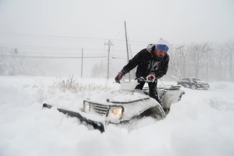 A man in heavy winter clothes rides an ATV through deep snow.