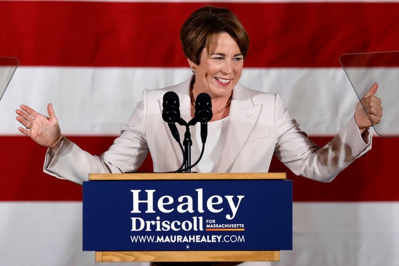 Individual with short dark hair wearing a white blazer, smiling, with arms up in victory at a podium.