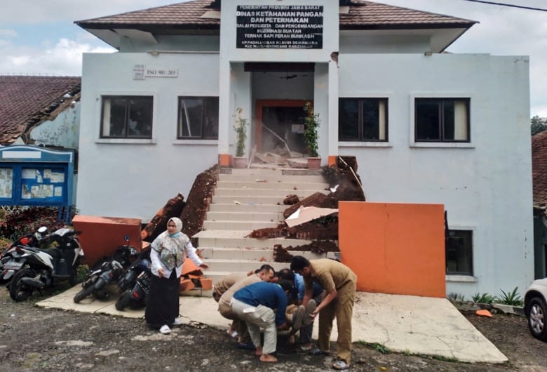 Four people help lift an unseen person in front of a damaged building.