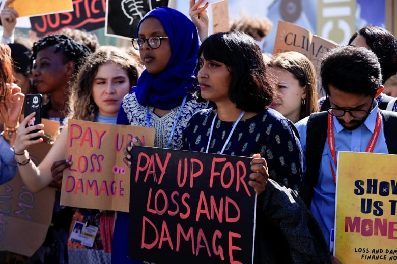 A young woman with shoulder-length brown hair and a nose ring stands at the front of a crowd of protesters, holding a sign that reads: "Pay up for loss and damage."