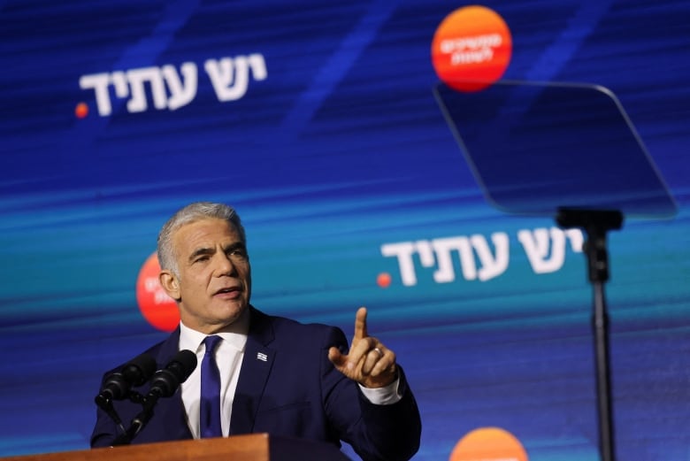 A grey haired individual wearing a blue suit and tie atanding at a podium, speaking with one arm raised. 