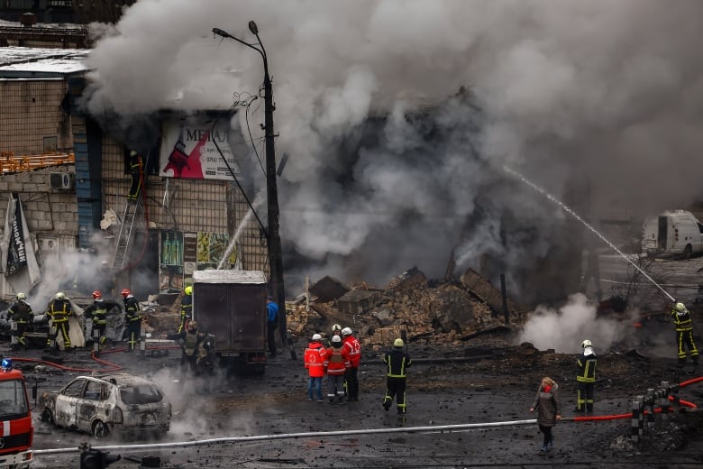 Firefighters spray water at a building surrounded by billowing black smoke. There's a large pile of rubble in front of the building, and a charred car in the street. 