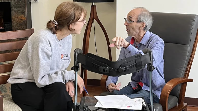 A woman sits next to an elderly man in a wheelchair. They are talking, the woman is smiling.