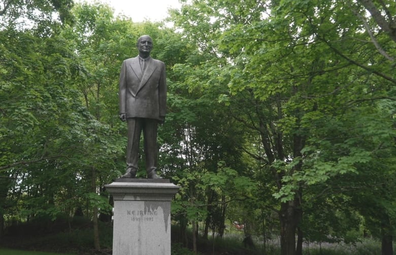 A statue of a man wearing a business suit is atop a pedestal in a park-like setting. The statue is surrounded by green foliage. 