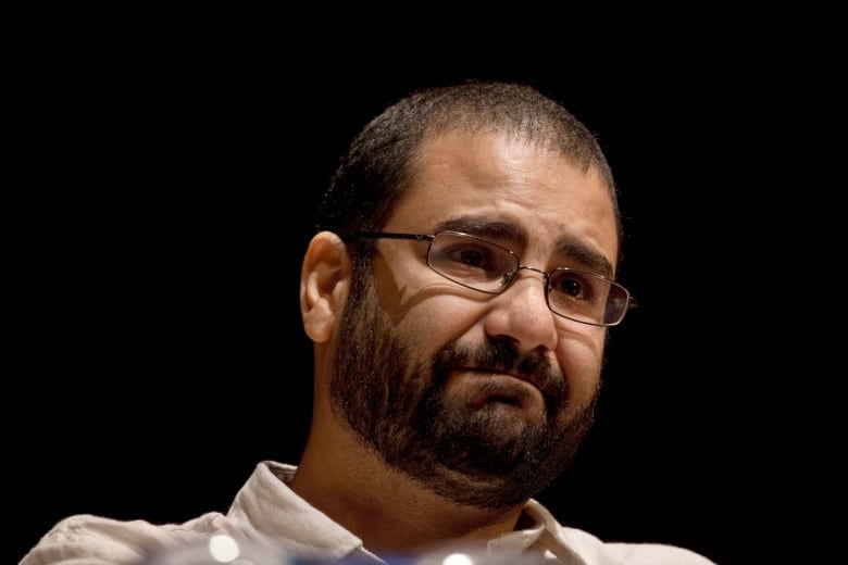 A frowning man with glasses and a black beard pictured from the shoulders up against a black backdrop. 