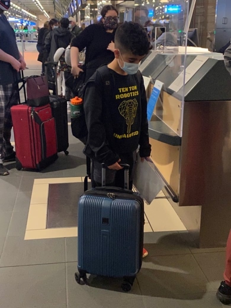 A masked boy tilts down his head while holding a trolly bag.