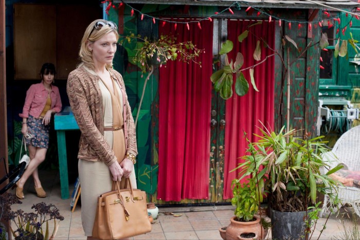 A smartly dressed blonde woman stands holding a handbag. She has a forlorn expression