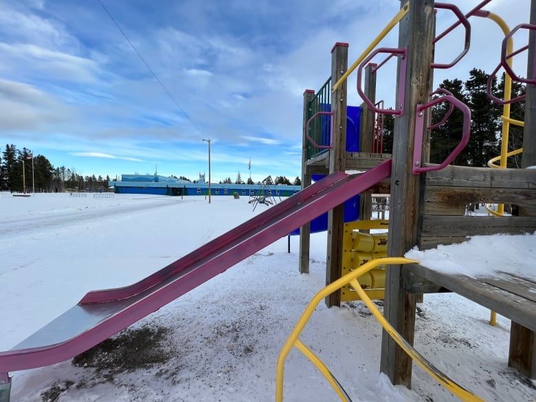 Colourful playground equipment, including a wide slide, is shown in the foreground of this image with a low-lying elementary school building painted in two tones of blue in the background.