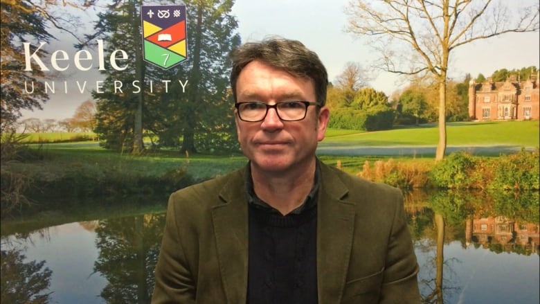 A man with short  hair, glasses and a green cord blazer sits in front of a branded Keele University backdrop with trees, a pond, buildings and the school logo. 