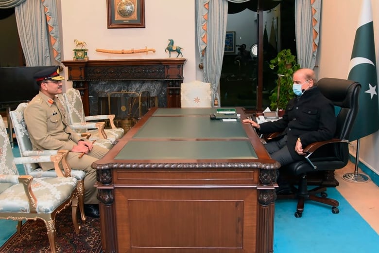 A man with glasses and a moustache wearing a military uniform sits across a desk from a balding man wearing a face mask. 