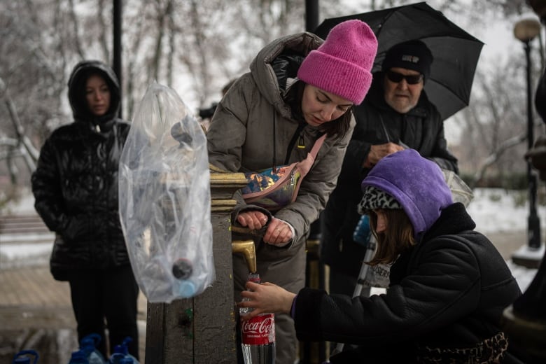 Individuals in winter coats and hats fill an empty Coke bottle at a public tap. 