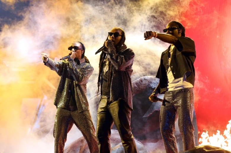 Three young men with long black dreadlocks, dark sunglasses and silver sequined outfits stand side-by-side on a stage with microphones as smoke, lit orange and red, billows behind them.