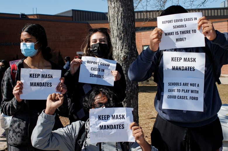 A group of high school-aged students wearing masks hold up signs that say "keep mask mandates" and "schools are not a game to play with COVID cases, Ford" and "Report COVID cases in schools"