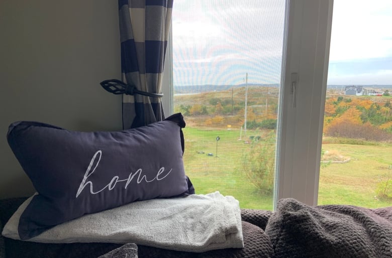 A pillow embroidered with the word "home" in cursive sits in front of a window. Trees with leaves turned Autumn colours can be seen through the window.