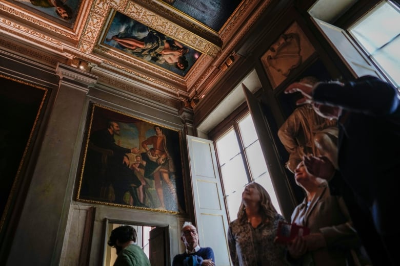 A group of people in a an art museum, photographed from below, stand beneath painting displayed on the ceiling. One man points towards it as two woman stare up and smile. Another man off to the side watches with arms folded. The painting shows a nude woman, draped in a long piece of fabric, sitting on clouds and holding a pot. 