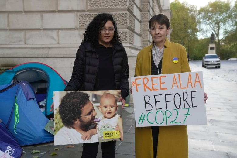 Two women stand side-by-side outside. One is holding photo of a man holding a smiling baby, with the Amnesty International logo in the corner. The other is holding a sign that reads: '#FreeAlaa before #COP27." Behind them is an unzipped blue tent, erected against a stone building. 