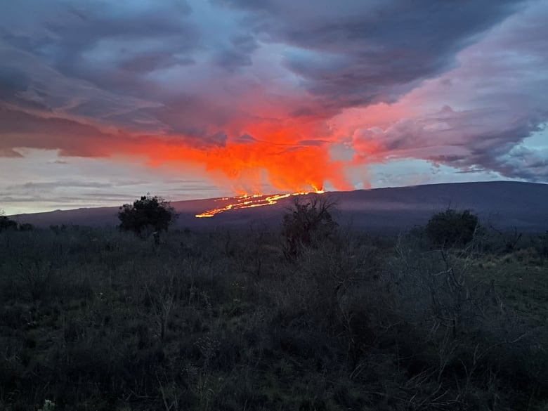 A distant view of a volcano with burning lava flowing out and clouds overhead.