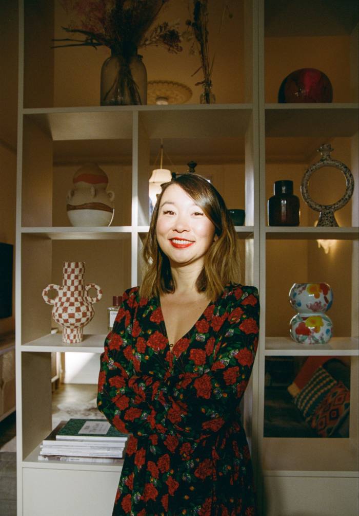 An Asian woman stands in front of a shelf unit that displays pieces of porcelain