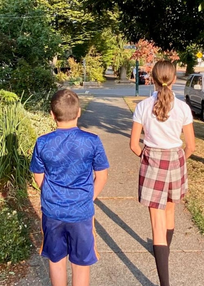 A girl in a uniform and a boy in regular clothes walk on a sidewalk with their backs to the camera.  