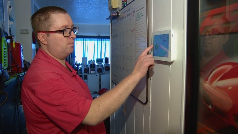 A white man wearing glasses and a red polo shirt touches a small security screen that is mounted on a white wall between a whiteboard calendar and a poster of a red sports car.