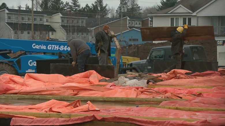 Orange fabric and slats of wood. Three men working in the background next to a piece of heavy machinery and a truck.