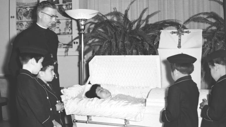 Photo shows priest and several children surrounding casket of deceased child. 